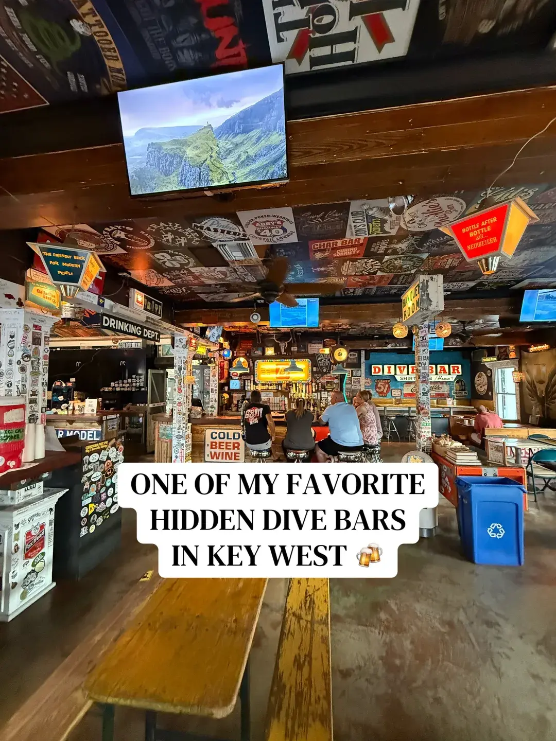 Interior of The Dive Bar Shirt Club in Key West, with a TV, neon 'Dive Bar' sign, and patrons seated at the bar under a sticker-covered ceiling