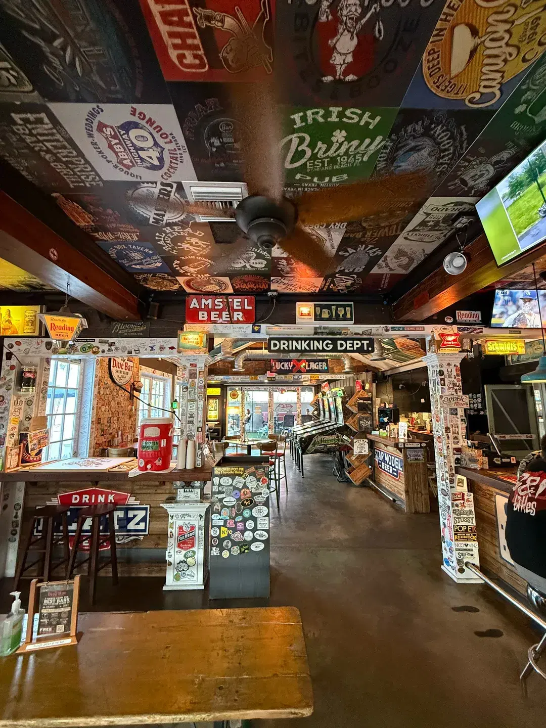 Hallway inside The Dive Bar Shirt Club with sticker-covered support columns, a 'Drinking Dept.' sign, and arcade games